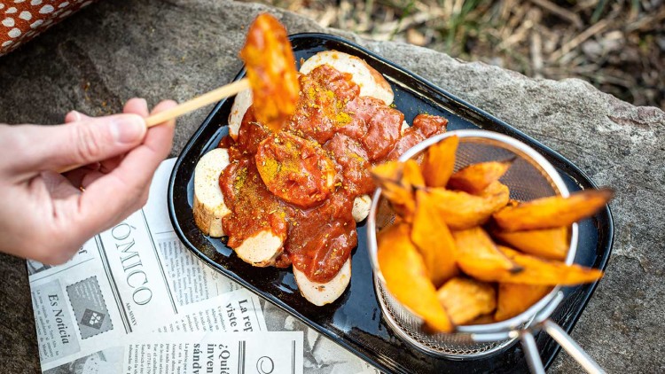 Schale mit Currywurst und Soße auf Baguettescheiben neben einem Sieb mit Süßkartoffelpommes auf Steinuntergrund.