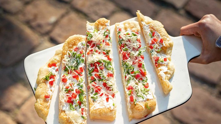 Geschnittene, belegte Teigfladen mit Creme, Paprika, Frühlingszwiebeln und Käse auf einem weißen Tablett, gehalten von zwei Händen.