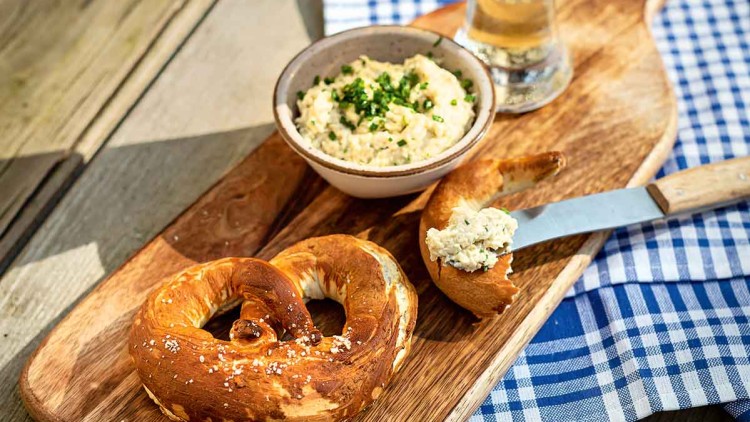 Eine Bretzel, Obazda mit Schnittlauch, ein Messer und ein Weizenglas stehen auf einem Holzbrett mit blau-weißem Tuch.