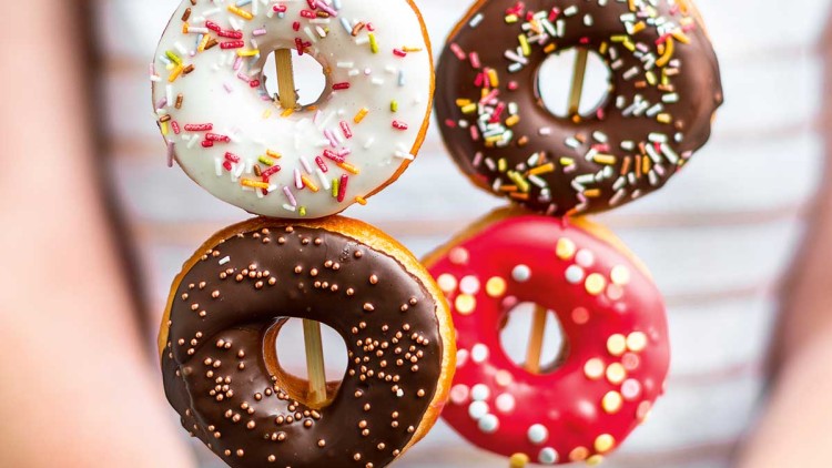 Sechs bunte Donuts mit Streuseln und Schokoglasur sind auf Holzstäben gesteckt und werden in Händen gehalten.