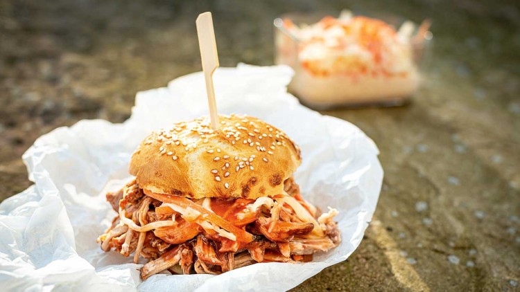Burger mit Pulled Pork, Krautsalat und Soße in einem Sesambrötchen, auf Backpapier serviert, im Hintergrund Krautsalat in einer Schale.