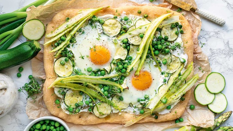 Flammkuchen mit zwei Spiegeleiern, grünem Spargel, Zucchinischeiben, Erbsen und frischen Kräutern auf Backpapier.