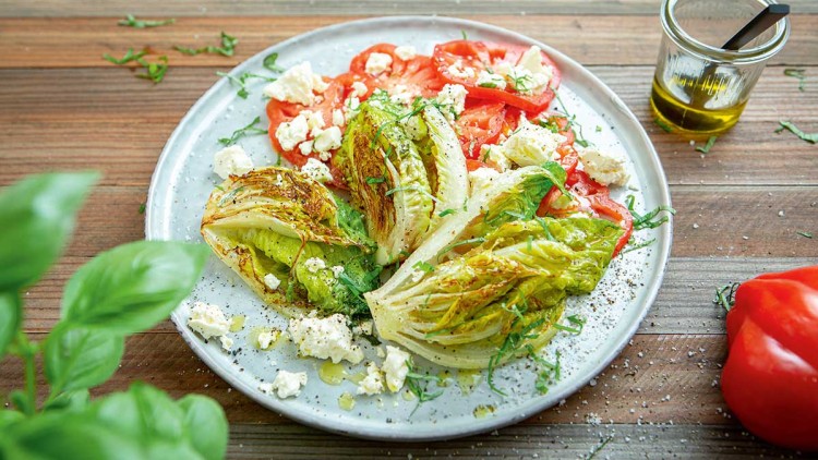 Gegrillter Salat mit Tomatenscheiben, zerbröseltem Feta, frischen Kräutern und Olivenöl auf einem weißen Teller.
