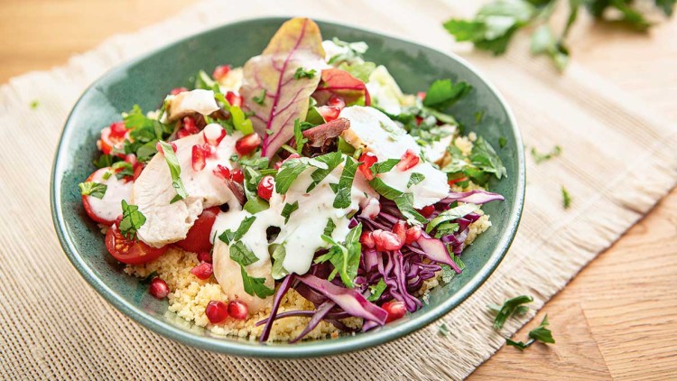 Bunter Salatteller mit Couscous, rotem Kraut, Granatapfelkernen, Tomaten, Kräutern und weißem Joghurt-Dressing.