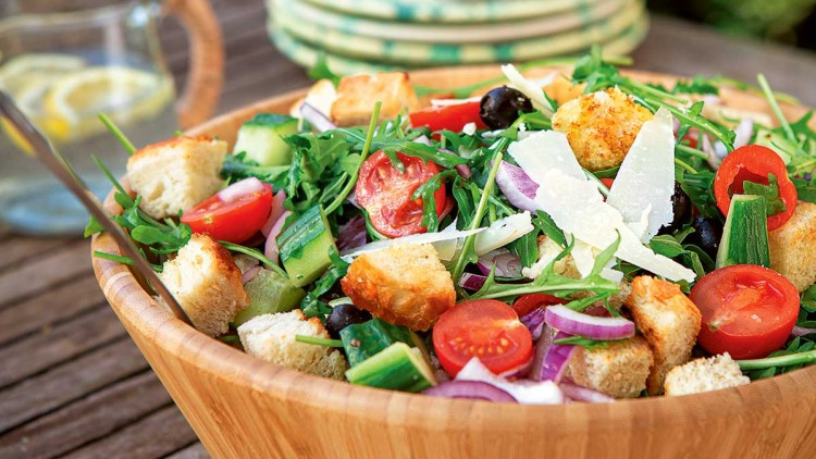 Große Holzschüssel mit gemischtem Salat aus Tomaten, Gurken, Rucola, Croutons, Oliven, Zwiebeln und Parmesan auf Holztisch.