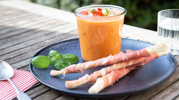 Ein Glas mit oranger Gazpacho, garniert mit Gemüse, auf einem Teller mit Basilikum und Schinkenstangen, daneben ein Wasserglas.