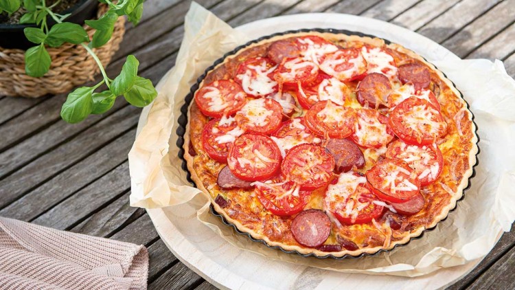 Frisch gebackene Quiche mit Tomatenscheiben, Salami und Käse auf Backpapier, Basilikumpflanze links im Hintergrund.