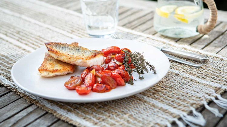 Ein Teller mit gebratenem Fisch, gerösteten Tomaten und Kräutern auf einem Holztisch mit Wasserglas und Karaffe im Hintergrund.