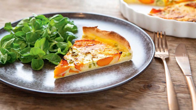 Ein Stück Kürbis-Quiche neben frischem Feldsalat auf einem schwarzen Teller, daneben liegen goldene Besteckteile auf Holztisch.