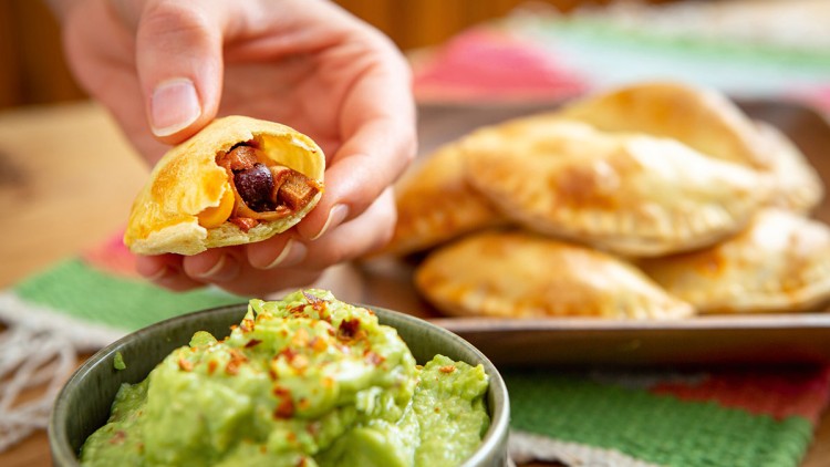 Hand hält gefüllte Teigtasche über grüne Guacamole in einer Schale, im Hintergrund weitere Teigtaschen auf einem Teller.