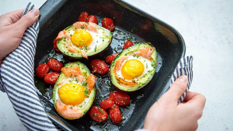 Avocadohälften mit Ei, Lachs und Kräutern in einer Auflaufform, umgeben von kleinen gebackenen Tomaten, werden gehalten.