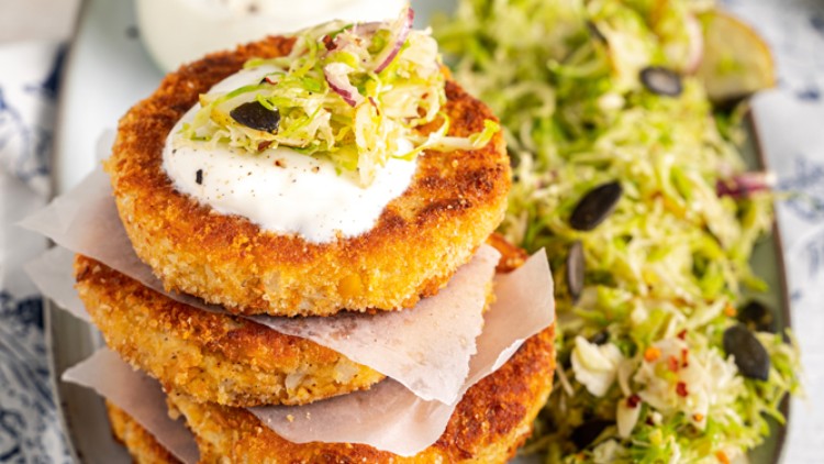 Goldbraune Gemüsepuffer mit Joghurt-Dip, Krautsalat und Kürbiskernen auf einem weißen Teller, daneben ein Glas mit Löffel.