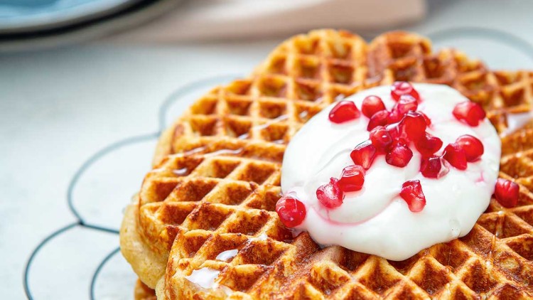 Frische goldbraune Waffeln mit Sahne und roten Beeren auf einem schwarzen Gitter, im Hintergrund ein blauer Teller.
