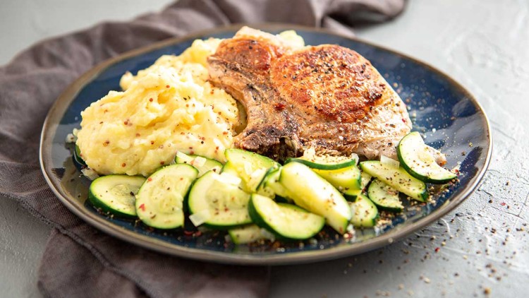 Ein gebratenes Kotelett mit Kartoffelpüree und gewürzten Zucchinischeiben auf einem dunklen Teller, umgeben von einem Stofftuch.