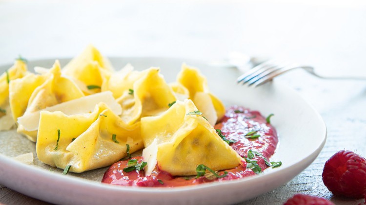 Gelbe, gefüllte Teigtaschen mit Kräutern auf roter Soße in einem weißen Teller, daneben frische Himbeeren und eine Gabel.