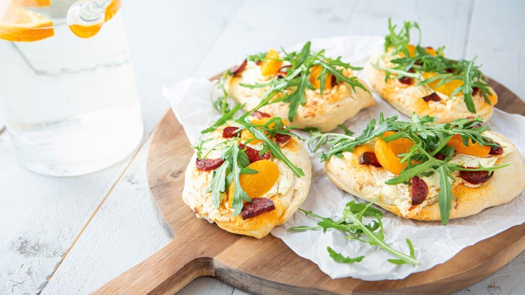 Vier kleine Pizzen mit Frischkäse, Mandarinen, Rucola und Wurst auf einem runden Holzbrett, links ein Glas mit Wasser.