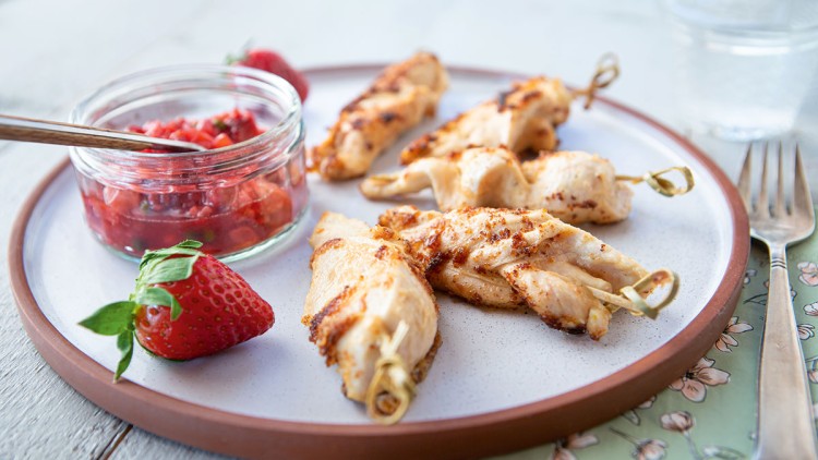Gegrillte Hähnchenspieße liegen auf einem Teller mit Erdbeeren und einem Glas mit roter Fruchtsoße.