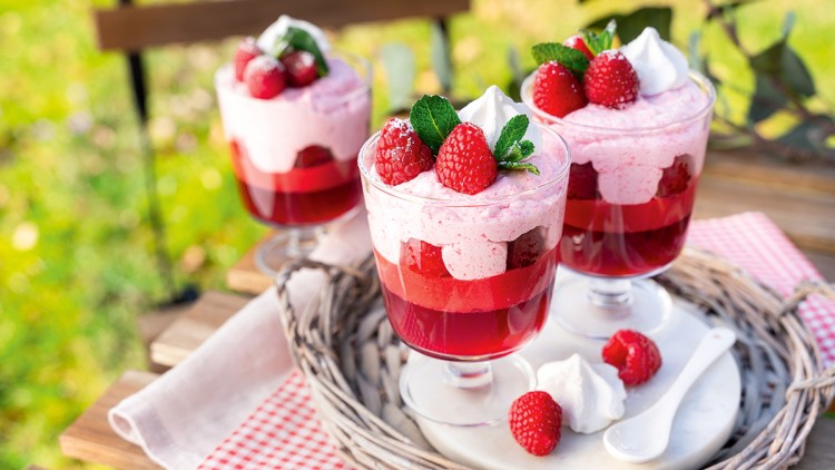 Drei Dessertgläser mit Himbeergelee, Creme, frischen Himbeeren, Minzblättern und Sahne auf einem Tablett im Freien.