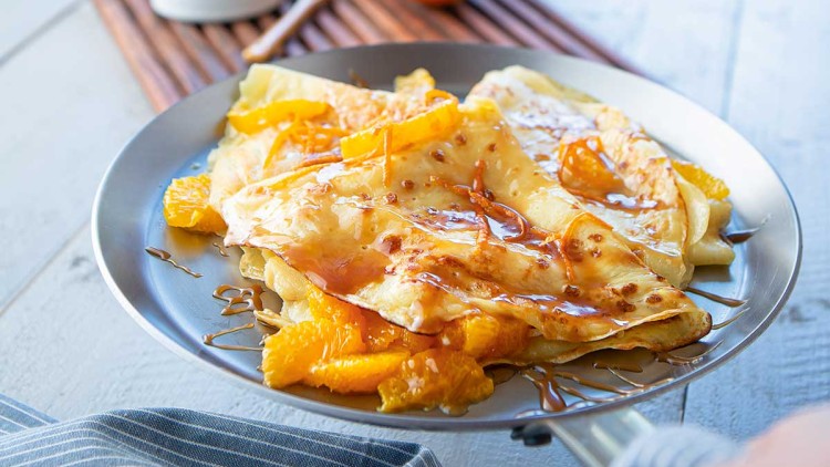 Crêpes mit Orangenspalten und Karamellsoße auf einem Teller, im Hintergrund Blumen, Mandarinen und ein Fenster.