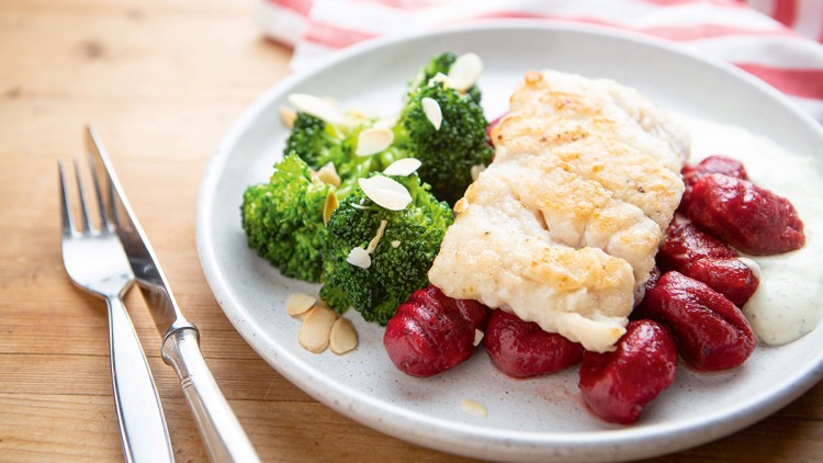 Gebratener Fisch mit Brokkoli, Rote-Bete-Gnocchi und heller Soße, dekoriert mit Mandelblättchen, auf einem hellen Teller.