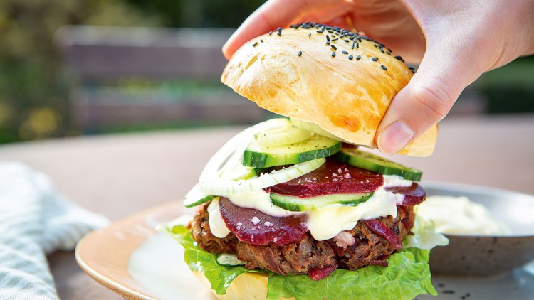 Hand hält einen Burger mit Salat, Gemüsepatty, Rote-Bete-Scheiben, Gurke, Zwiebel, Sauce und Sesam-Brötchen auf einem Teller.