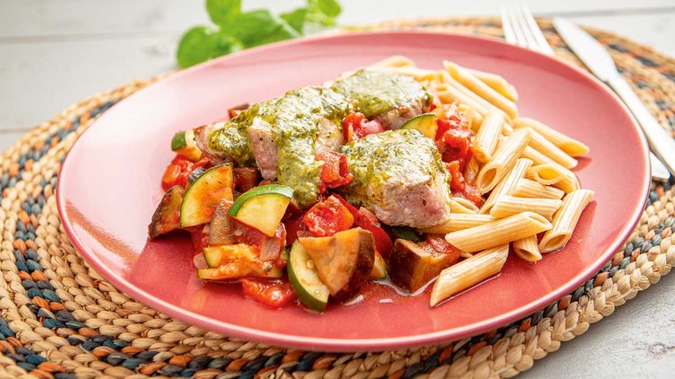 Rosa Teller mit Penne-Nudeln, Gemüse, Fleischstücken mit grünem Pesto, auf geflochtenem Untersetzer mit Besteck.