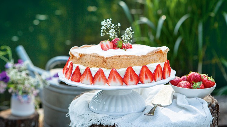 Erdbeerkuchen mit Sahne und Deko aus kleinen weißen Blüten steht auf weißer Tortenplatte neben Schale mit Erdbeeren.