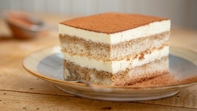 Ein Stück Tiramisu mit Kakaopulver bedeckt liegt auf einem Teller auf einem Holztisch, im Hintergrund ein Sieb und eine Mokkakanne.