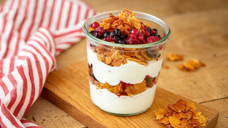 Schichtdessert aus Joghurt, Beeren und karamellisierten Mandeln in einem Glas auf einem Holzbrett, daneben ein gestreiftes Tuch.