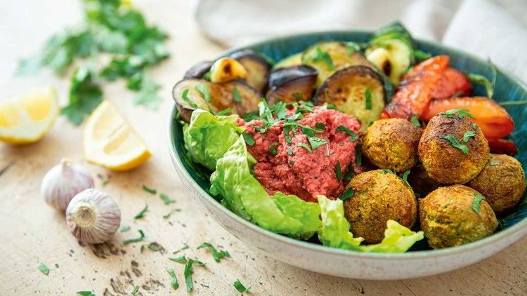 Bunte Schale mit Falafel, Rote-Bete-Dip, gegrilltem Gemüse und frischem Salat, im Hintergrund Knoblauch, Zitrone und Kräuter.