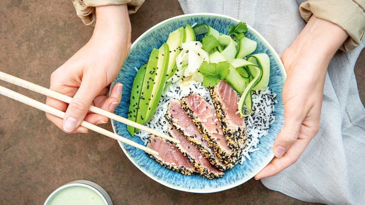 Eine blau-weiße Schale mit Reis, Avocado, Gurke, Sesam und in Scheiben geschnittenem Thunfisch wird mit Stäbchen gehalten.