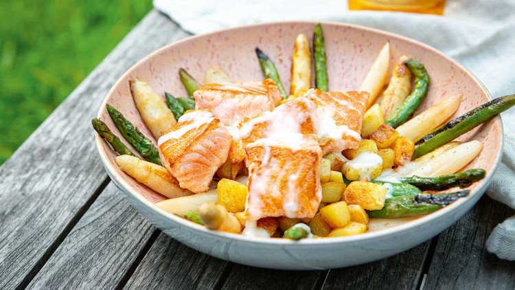 Gegrillte Lachswürfel mit weißem und grünem Spargel, Kartoffelwürfeln und Soße in einer Schale auf einem Holztisch.