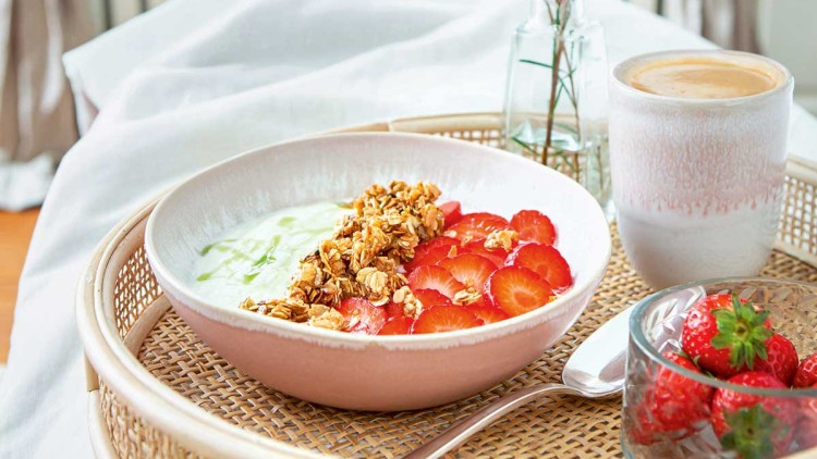 Eine Schale mit Joghurt, Granola und Erdbeerscheiben auf einem Tablett, daneben ein Glas Kaffee, Erdbeeren und eine Vase mit rosa Blumen.