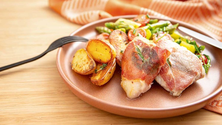 Ein Teller mit gebratenen Kartoffeln, grünem Spargel, Kirschtomaten und in Schinken gewickeltem Fisch, daneben Gabel und Messer.