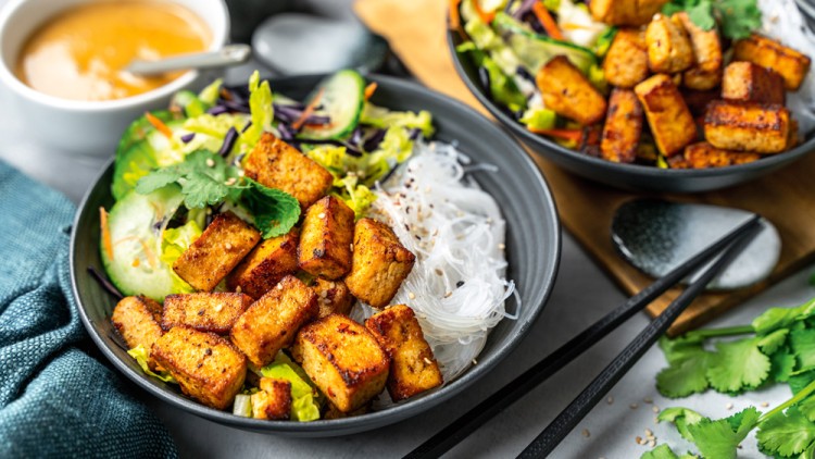 Schwarze Schale mit gebratenem Tofu, Glasnudeln, Gurkenscheiben, Salat und Kräutern, daneben ein Dip in einer weißen Schale.