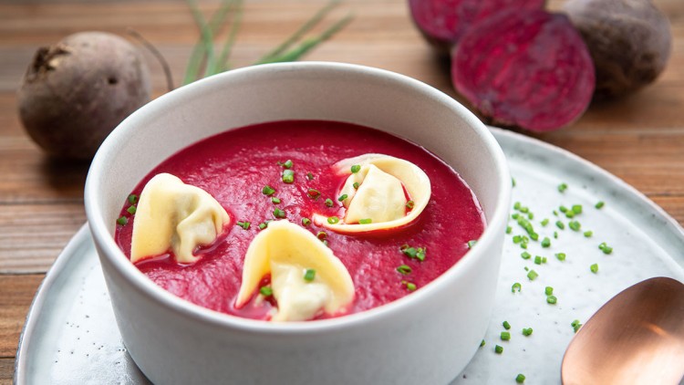 Eine weiße Schale mit roter Rote-Bete-Suppe, gefüllten Teigtaschen und Schnittlauchringen auf einem Teller mit Kupferlöffel.