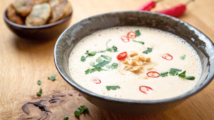 Cremige Suppe in grauer Schale mit ganzen Erdnüssen, Petersilie und roten Chiliringe, daneben braune Schüssel mit Gebäck.