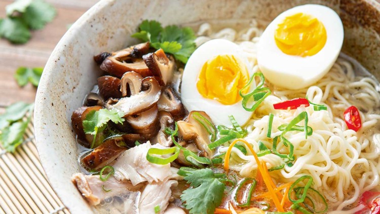 Keramikschale mit Ramen-Nudeln, Ei, Pilzen, Karotten, Chili und Kräutern, daneben Holzschale mit roten Chilischeiben.