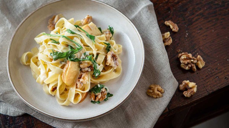 Tagliatelle mit Rucola, Walnüssen und Sahnesoße auf einem weißen Teller, daneben Walnüsse auf einem Holztisch.