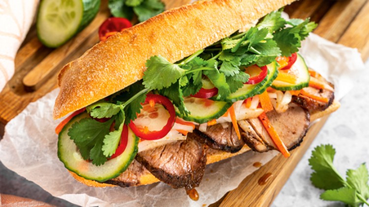Zwei belegte Baguettes mit Fleisch, Gurkenscheiben, Karotten, Paprika und Koriander auf Holzbrett, daneben Soße in einer Schale.