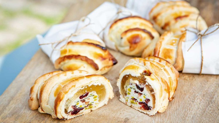 Blätterteiggebäck gefüllt mit Frischkäse, getrockneten Cranberrys und Pistazien liegt auf einem Holzbrett.