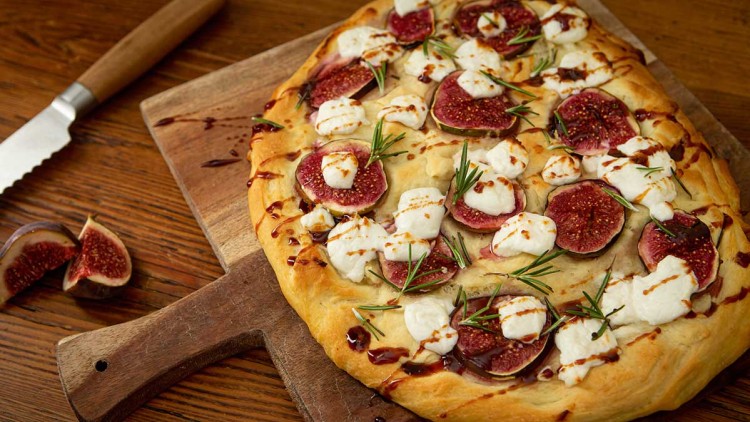 Flammkuchen mit frischen Feigen, Ziegenkäse, Rosmarin und Balsamico-Glasur auf einem hölzernen Schneidebrett.