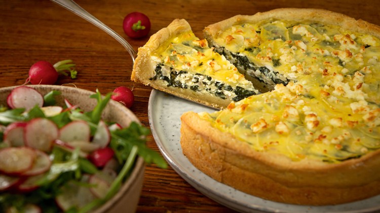 Ein Stück Käse-Spinat-Quiche wird von einem Metallheber herausgehoben, daneben steht eine Schüssel mit Radieschen-Rucola-Salat.