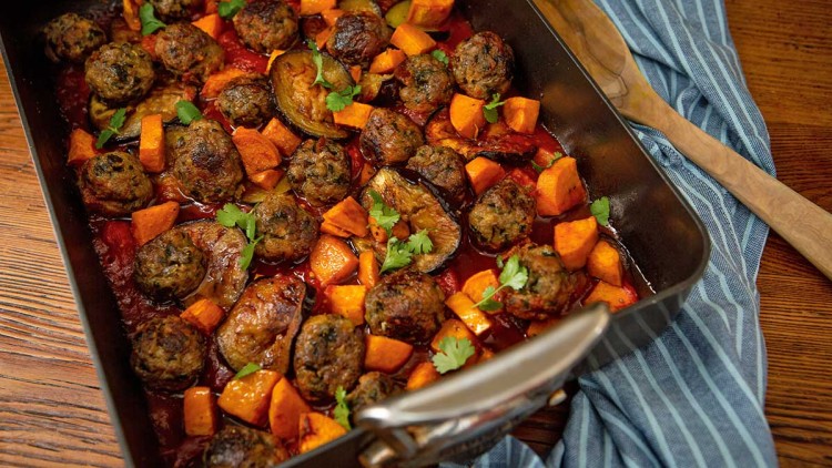 Ofenform mit Hackbällchen, Süßkartoffelstücken, Auberginenscheiben in Tomatensauce und frischen Kräutern auf Holztisch.