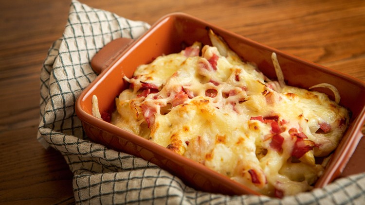 Überbackene Kartoffeln mit Käse und Schinkenstreifen in einer orangefarbenen Auflaufform auf einem karierten Küchentuch.