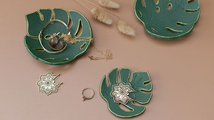 Grüne Schalen in Monstera-Blattform mit goldenem Schmuck und einem Ring auf rosa Untergrund, dekoriert mit Trockenblumen.
