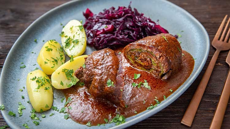 Ein Teller mit zwei Rinderrouladen in brauner Soße, Kartoffeln mit Petersilie und Rotkohl, daneben goldfarbenes Besteck.