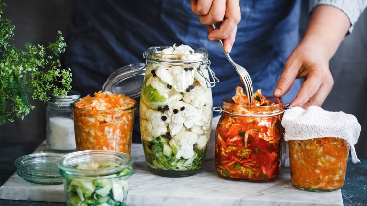 Verschiedene Glasbehälter mit eingelegtem Gemüse wie Blumenkohl, Karotten und Gurken stehen auf einer hellen Unterlage.