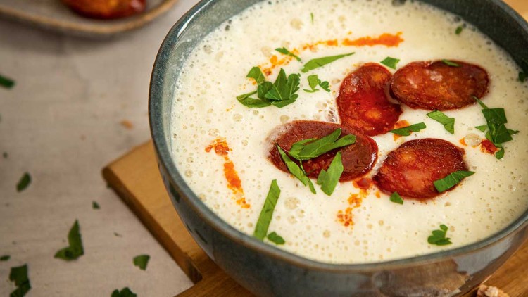 Cremige Suppe in einer Schale mit Chorizo-Scheiben und Petersilie, daneben Holzbrett mit Baguette und Teller mit Chorizo.