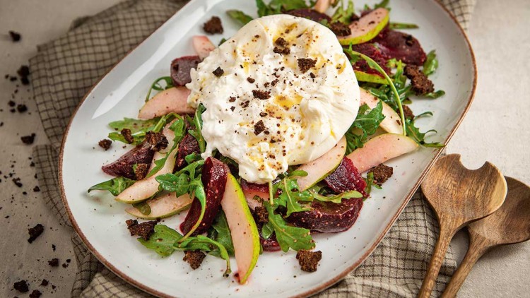 Ein Teller mit Salat aus Rucola, Birnenspalten, Roter Bete, Burrata, Brotstücken und Pfeffer, daneben hölzerne Salatbestecke.
